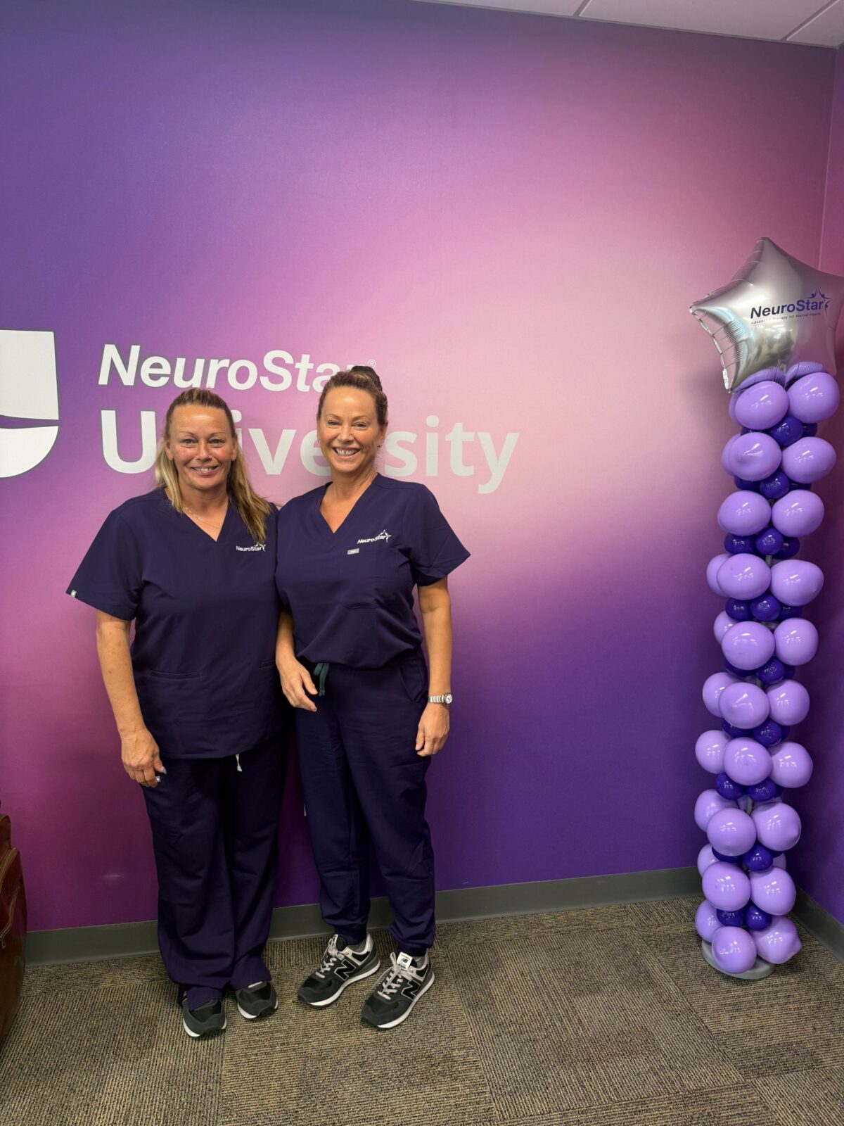 Two Mediterra Mindful Care team members in NeuroStar scrubs smiling in front of the NeuroStar University wall after completing their NeuroStar TMS training